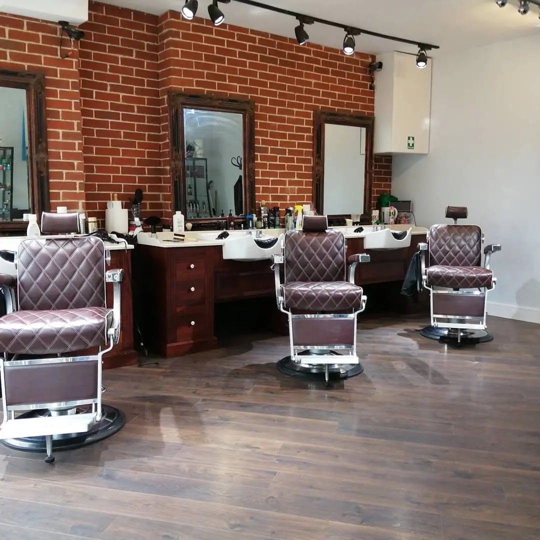 Inside Gentleman's Barber in Whitechapel: exposed red-brick wall, ornate dark-wood framed mirrors and classic brown leather barber chairs on a warm hardwood floor.